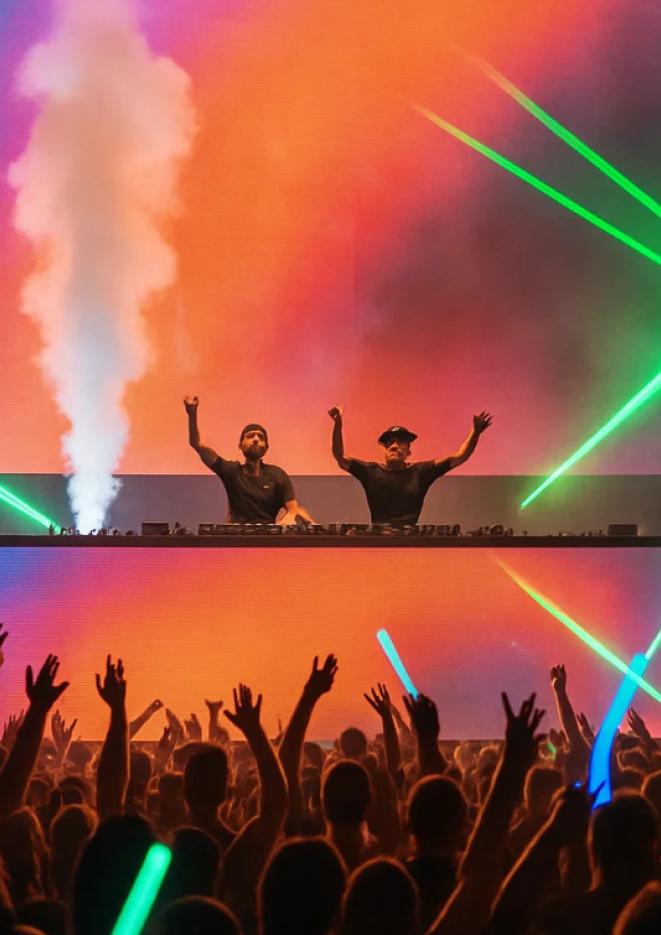 Two DJs on stage with their arms in the air, surrounded by orange light with greens strobes.