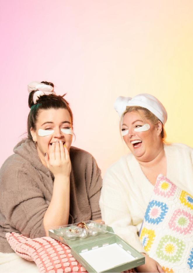 Female comedians Mel Moon and Tal Davies sit against a pastel pink and yellow background in their pyjamas eating chocolates and laughing.