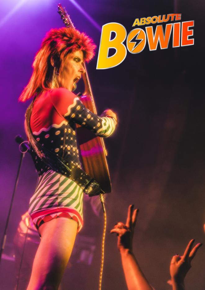 A David Bowie tribute artist performs on stage in a bright costume, holding up a guitar.