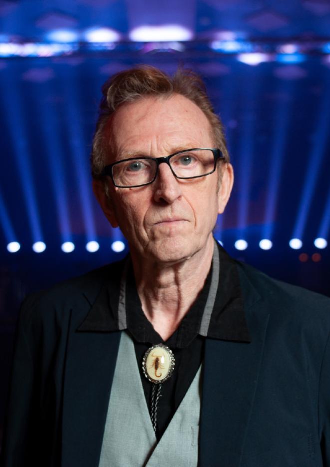 A close up of musician Bruce Watston wearing a suit, stood against a background with blue spotlights.