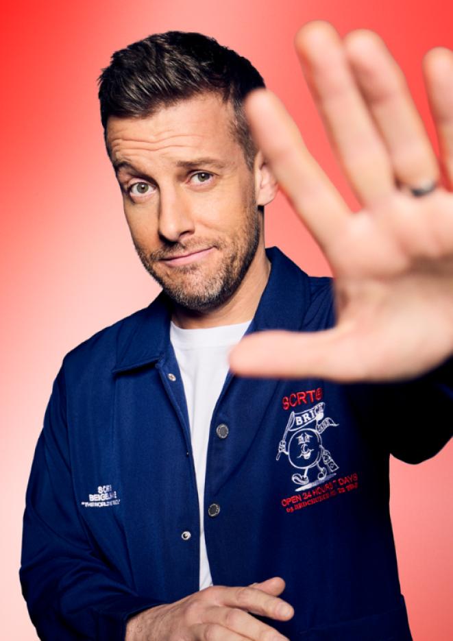 Chris Ramsey smiling with his hand reaching out to the camera, against a red background. He wears a white top with a blue jacket.