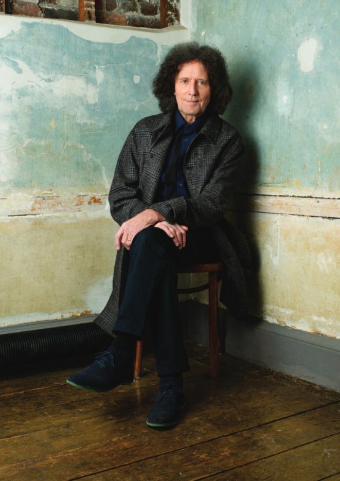 A man in a black coat sits on a chair in the corner of a room with blue wallpaper