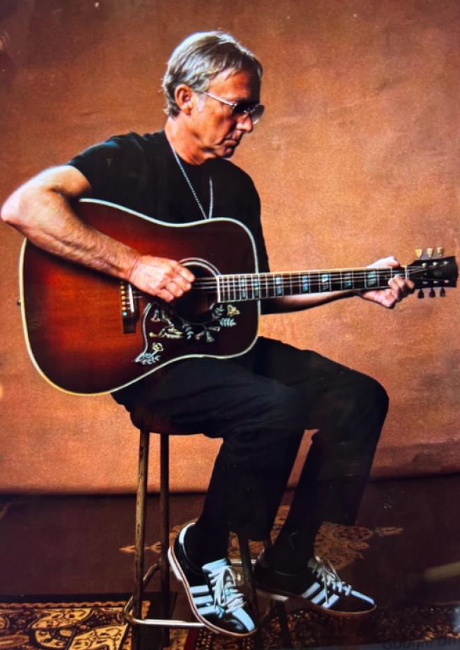 Guitarist Steve Craddock sits on a stool against an orange background, playing guitar.