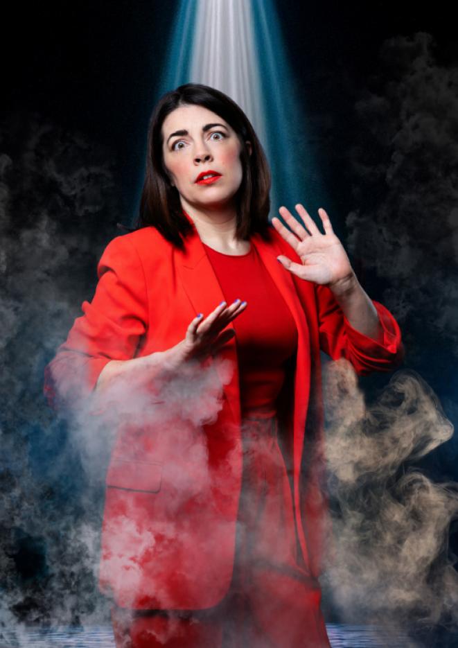 Comedian Rosie Holt wears a red suit against a black background with smoke in front of her and a light shining down.