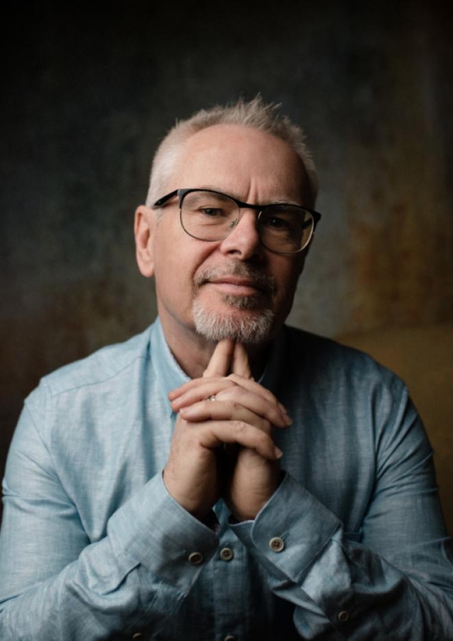 A close up image of musician Nik Kershaw sat against a dark backdrop, wearing a light blue shirt.
