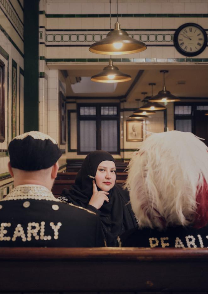 Fatiha El-Ghorri sits at a restaurant table with her head in her hand, smirking at the camera, opposite two people wearing black tops with 'Pearly' on the backs..