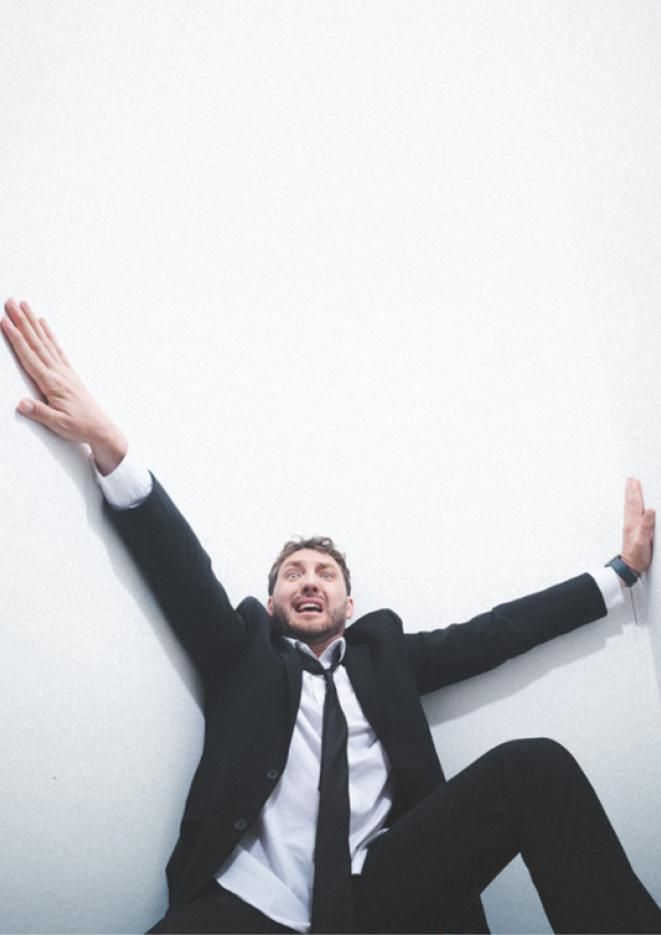 Comedian Seann Walsh wearing a black suit, clinging onto the walls of a small, white room.