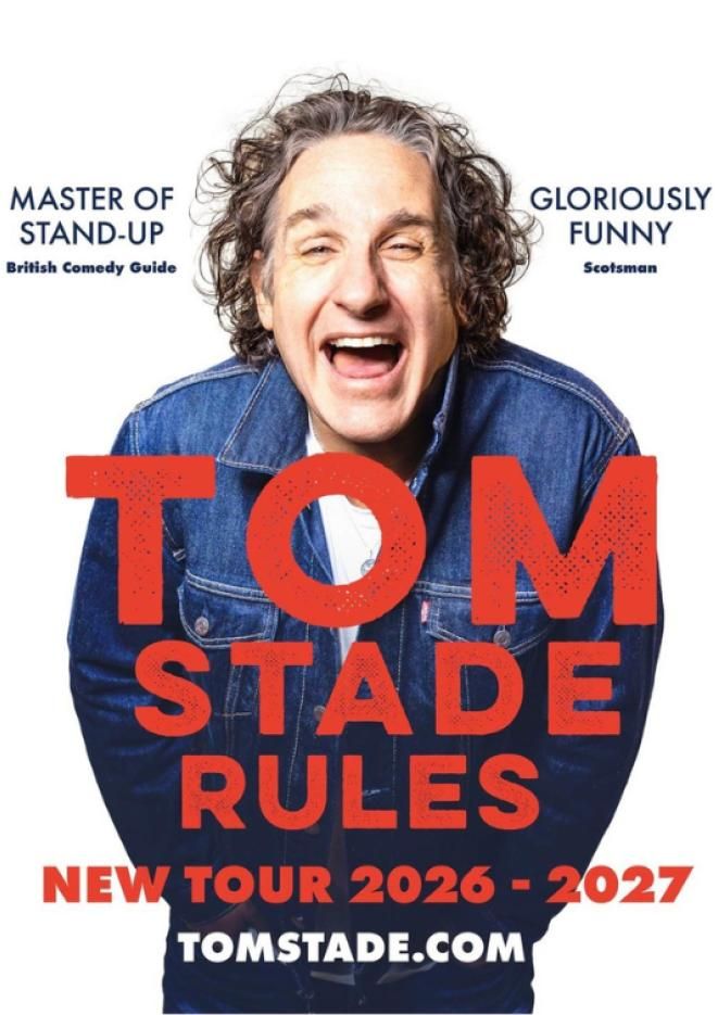 Comedian Tom Stade laughs, stood against a plain white background, wearing a blue denim jacket. Red text over the top reads Tom Stade Rules.