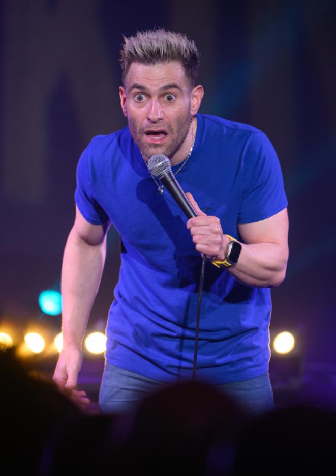 Comedian Simon Brodkin stands on stage holding a microphone, speaking to people in the audience.