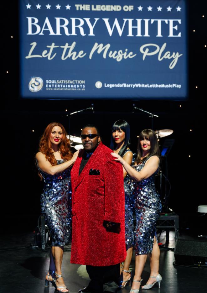A photo of singer William Hicks on stage, wearing a long red coat, with three women wearing blue sparkly dresses stood around him.