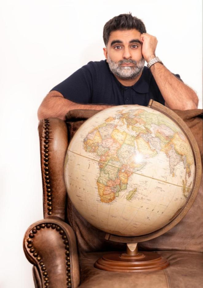 Comedian Tez Ilyas leans on a brown, leather armchair with a globe sat on it.