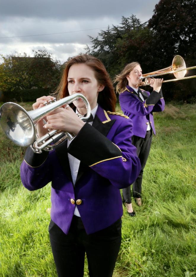 Two young women in purple blazers play brass instruments in a field.
