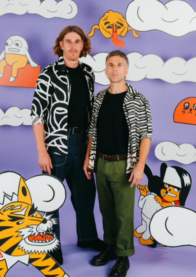 Two white men in black and white shirts stand in front of a purple background with cartoon clowns and creatures.