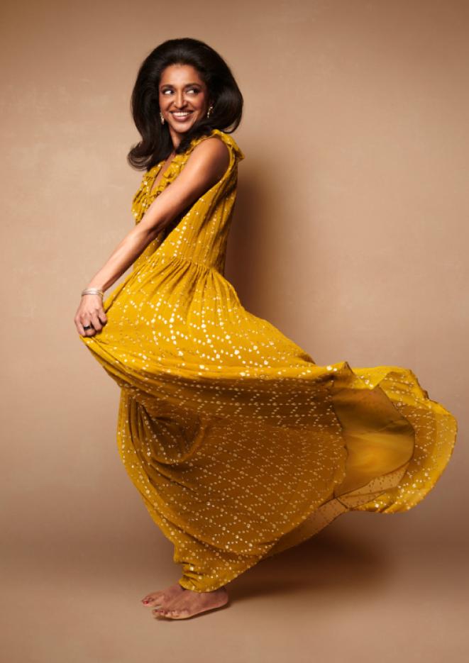 Comedian Sindhu Vee stands against a beige background, twirling in a long, yellow dress.