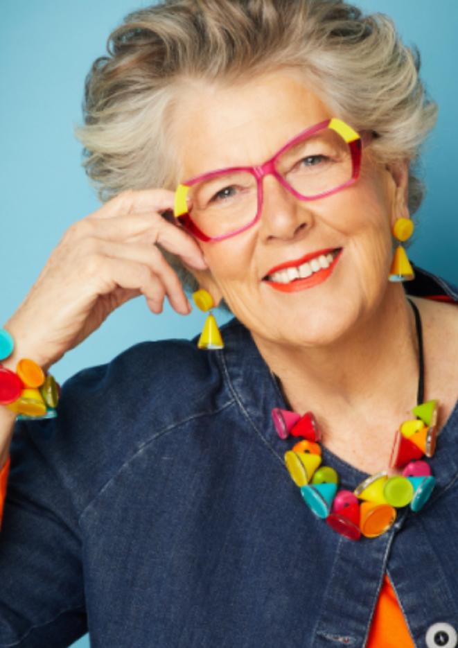 Prue Leith sits in front of a blue background wearing bright, multi-coloured jewellery.