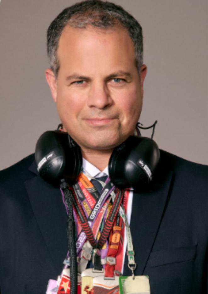 Motorsport commentator Ted Kravitz stands in front of a plain, grey background wearing headphones and multiple lanyards.