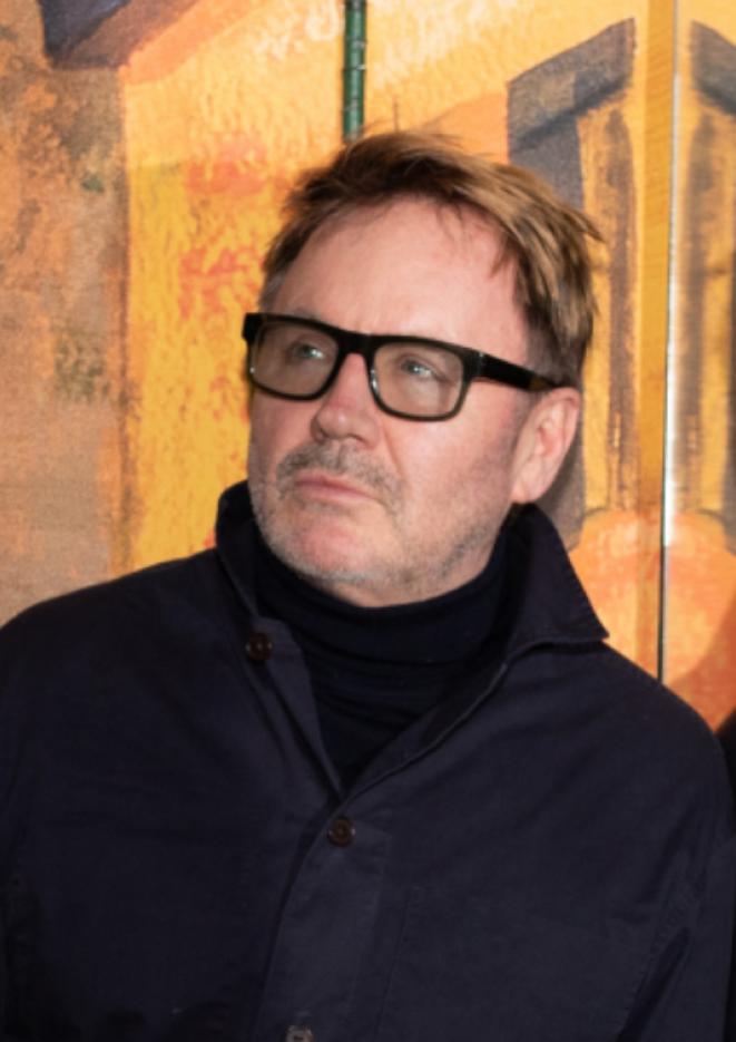 Artist and curator James Payne wears all black as he stands against an orange background.