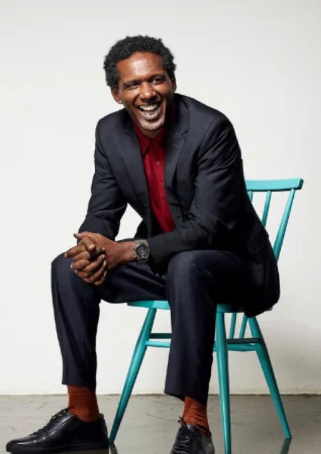 Lemn Sissay sits on a blue chair against a plain, white background, laughing to the camera.