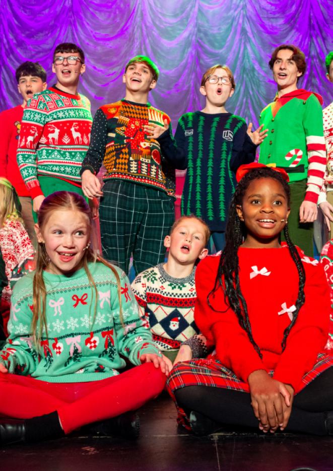 A group of school children wearing bright Christmas jumpers sing on stage.