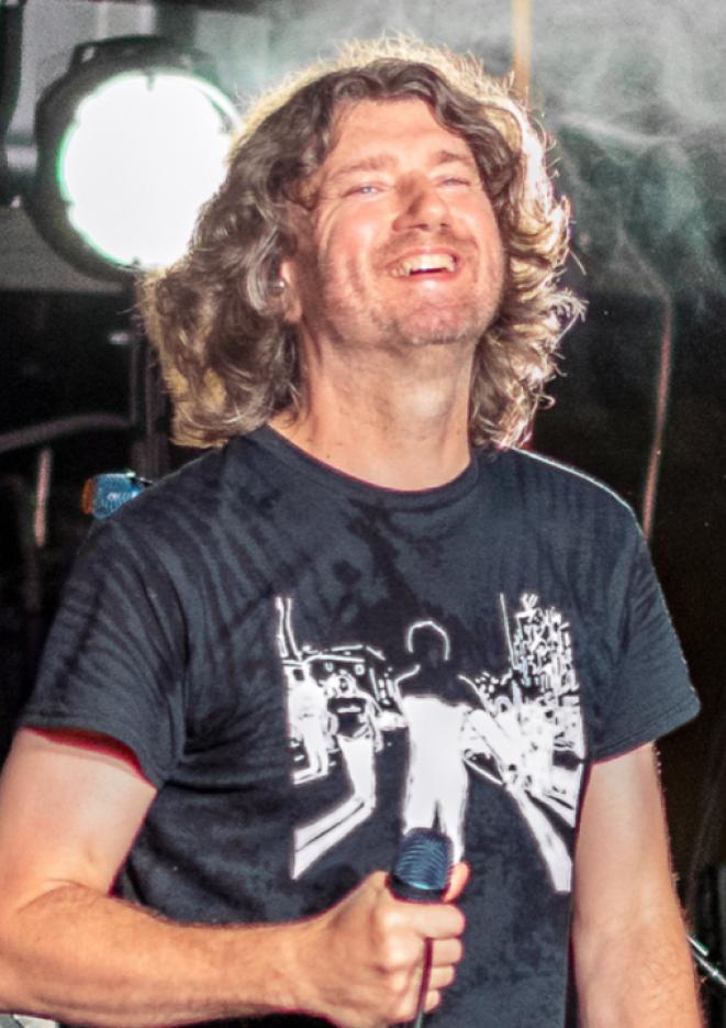 Musician Danny McNamara stands on stage holding a microphone. Dry ice swirls behind him.