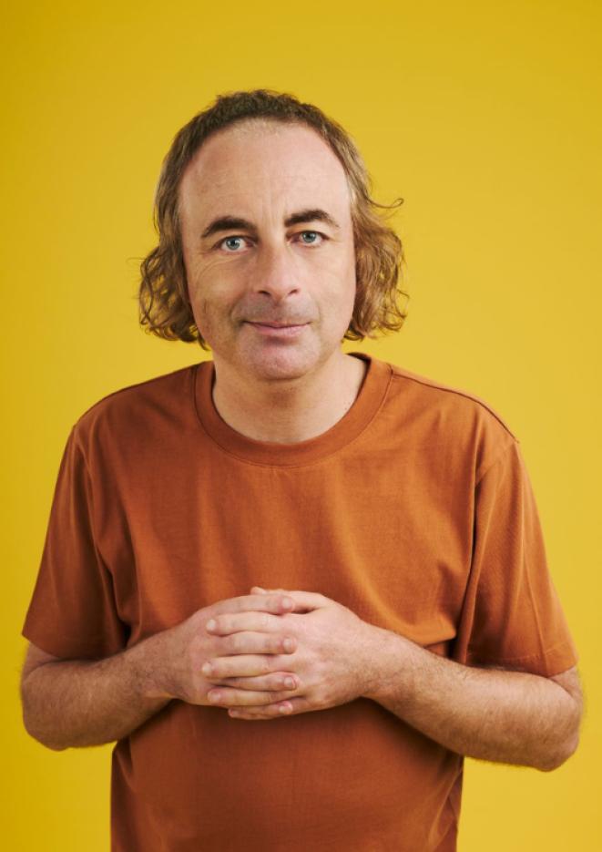 Comedian Paul Foot stands in front of a plain yellow background, wearing an orange tshirt.