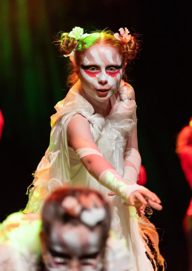 A young girl with red pigtails reaches out on a stage, with a white dress on and white and pink face paint.