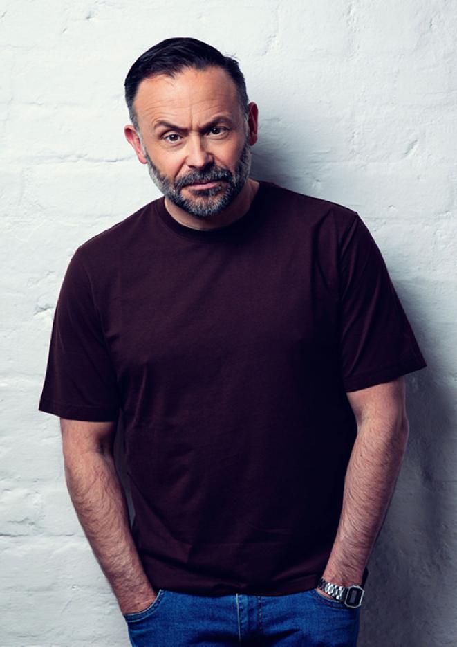 Comedian Geoff Norcott stands against a white brick wall with his hands in his jean pockets.
