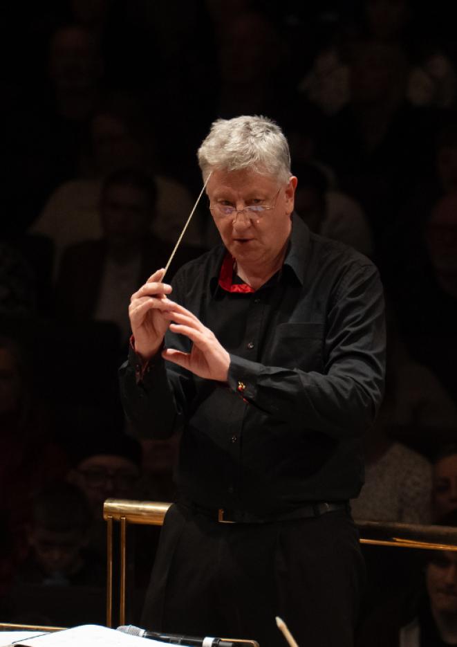 A man in a black suit leads an orchestra on stage
