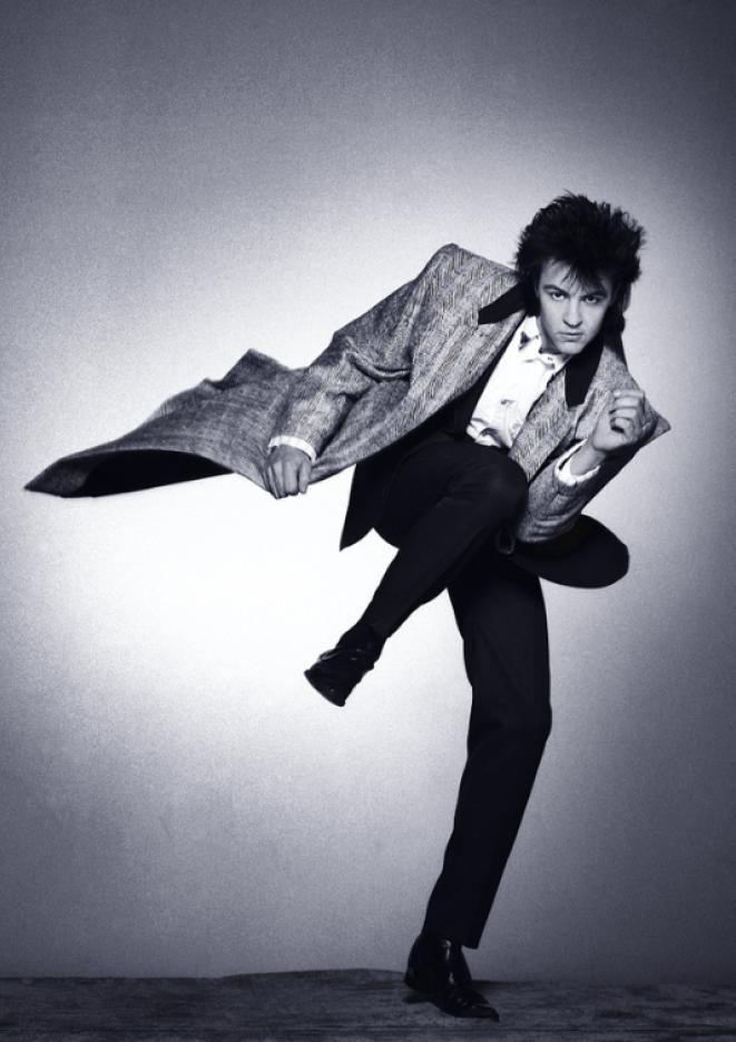 Black and white image of singer Paul Young kicking his leg in the air, wearing a suit and long jacket.