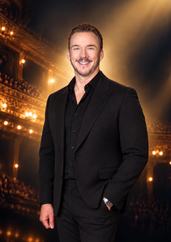A man in a black suit smiles at the camera in front of an auditorium