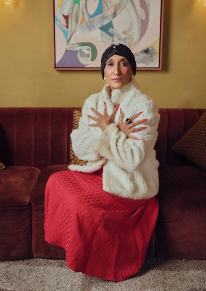 A woman in a red skirt, white fluffy coat and black headwear, sits on a chair with her hands on her chest