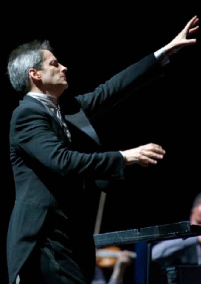 A man in a black suit conducts an orchestra against a black background