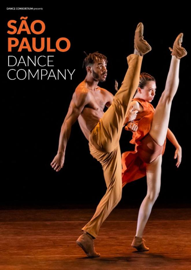 A male and female dancer wearing orange costumes lift their legs in the air in a synchronised move.