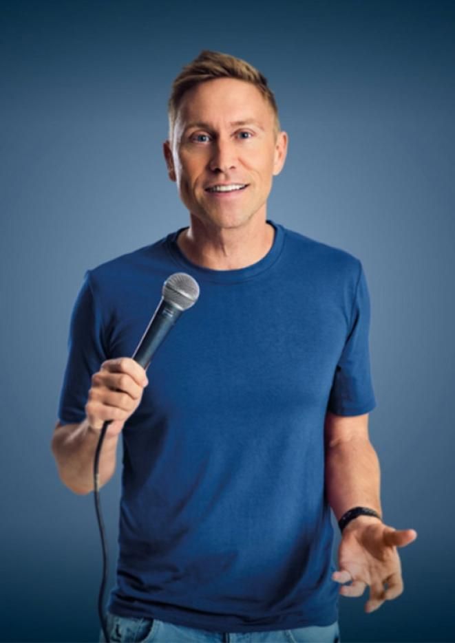 Comedian Russell Howard holds a microphone, stood against a blue background.